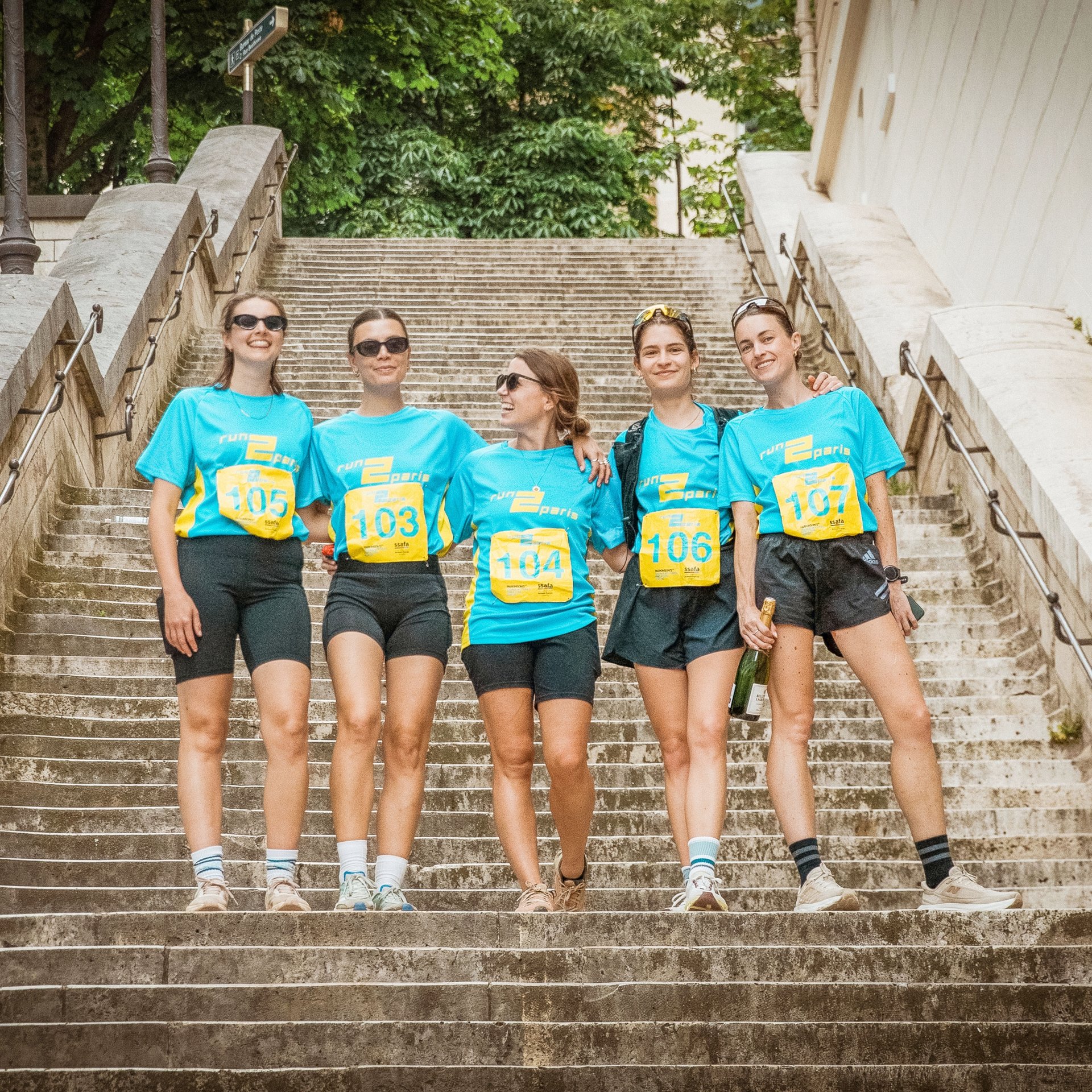 Bright female runners in blue event shirts standing on a staircase in Paris, celebrating during the Run2Paris relay race.
