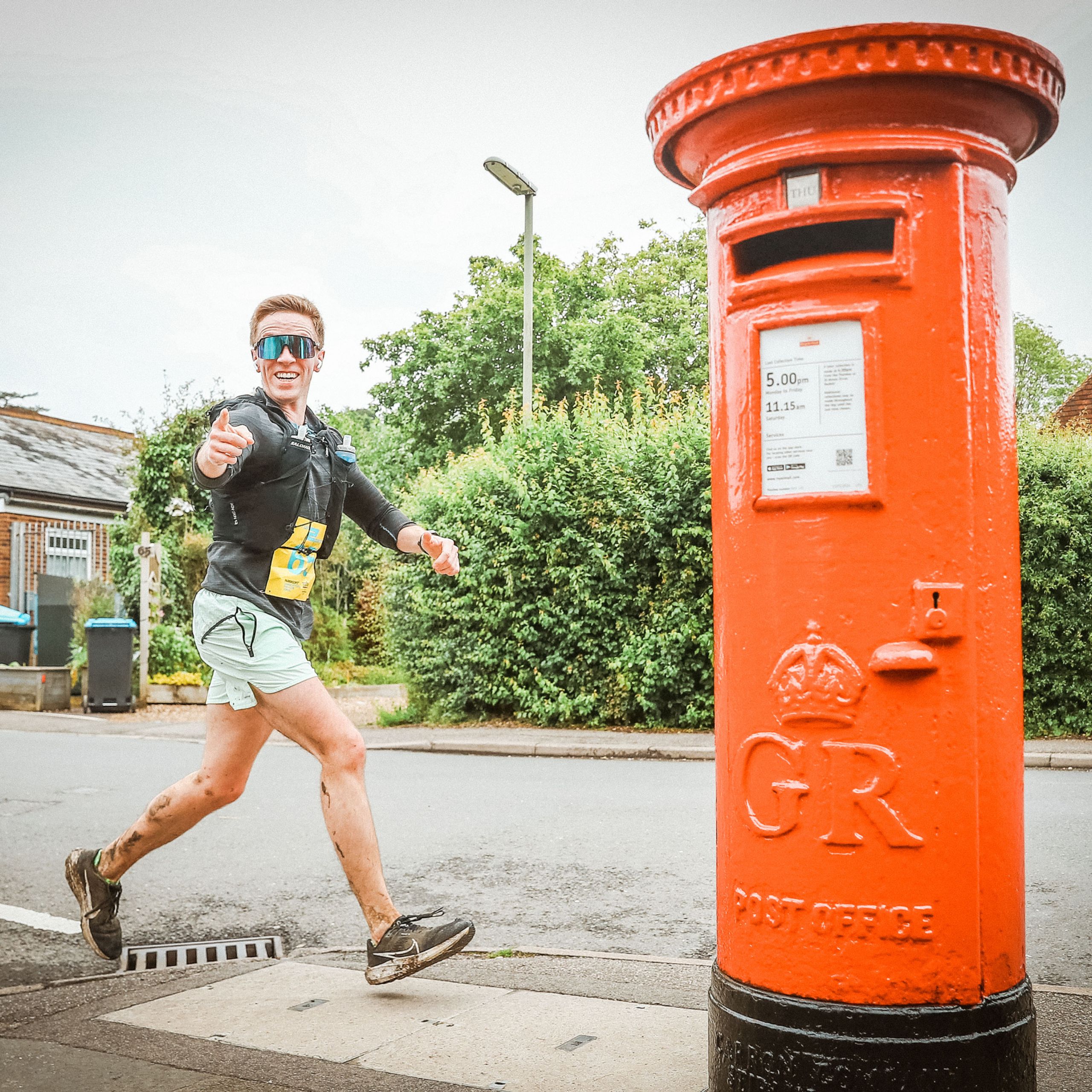 Bright male runner in sunglasses smiling near a traditional red post box during a relay race.