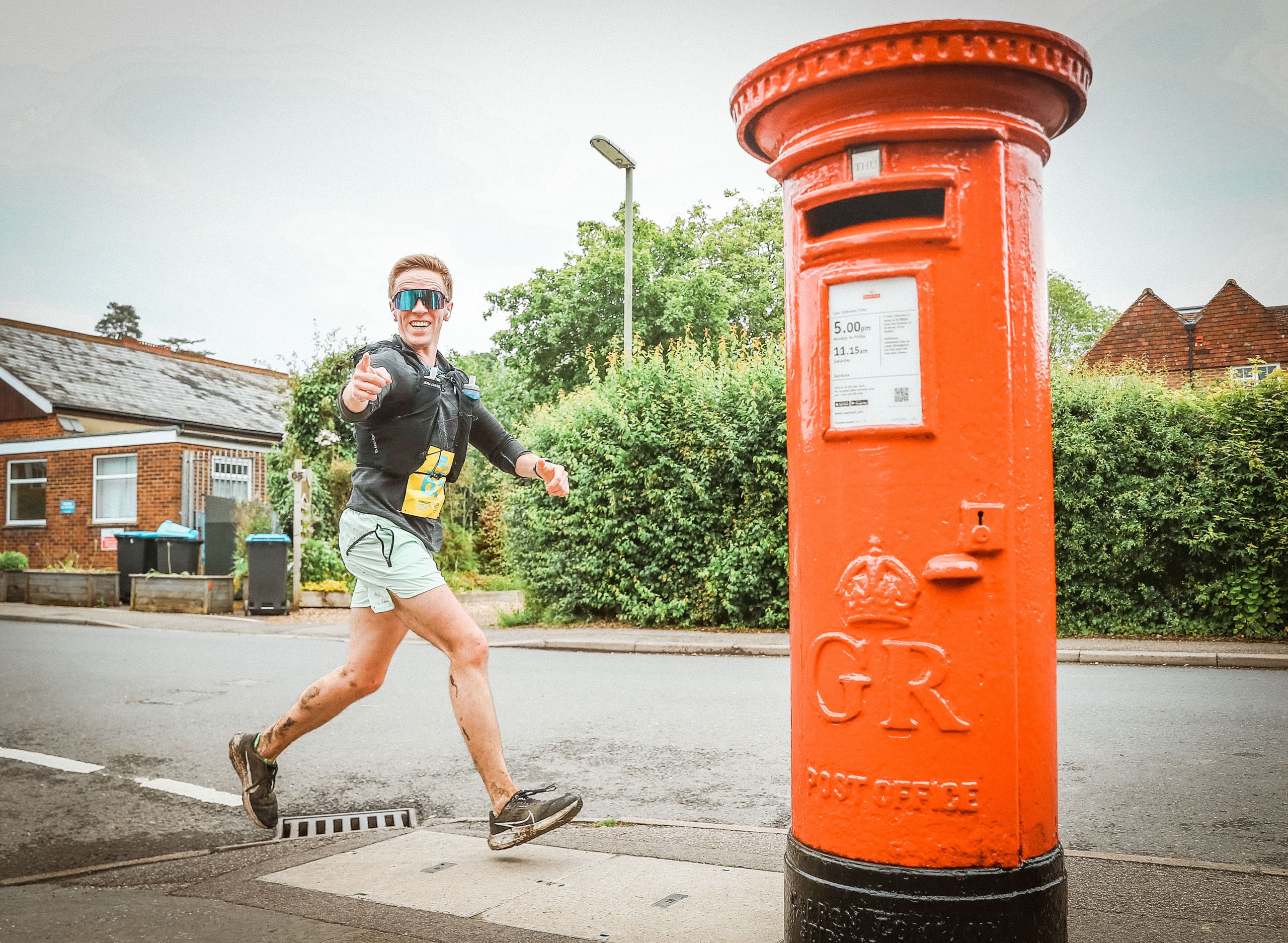 Bright male runner in sunglasses smiling near a traditional red post box during a relay race.