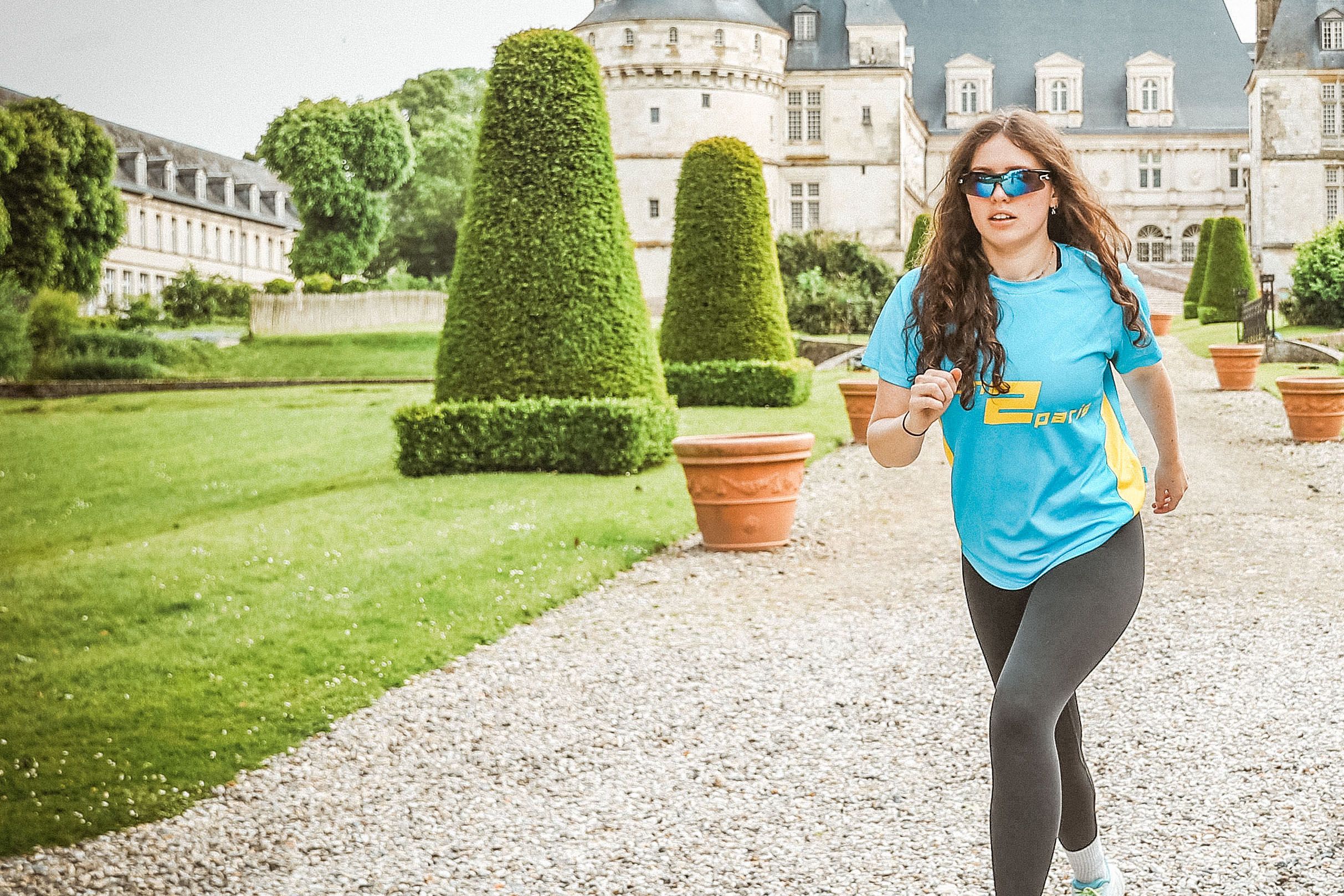 Running woman in blue running shirt, black leggings, and sunglasses in a garden.