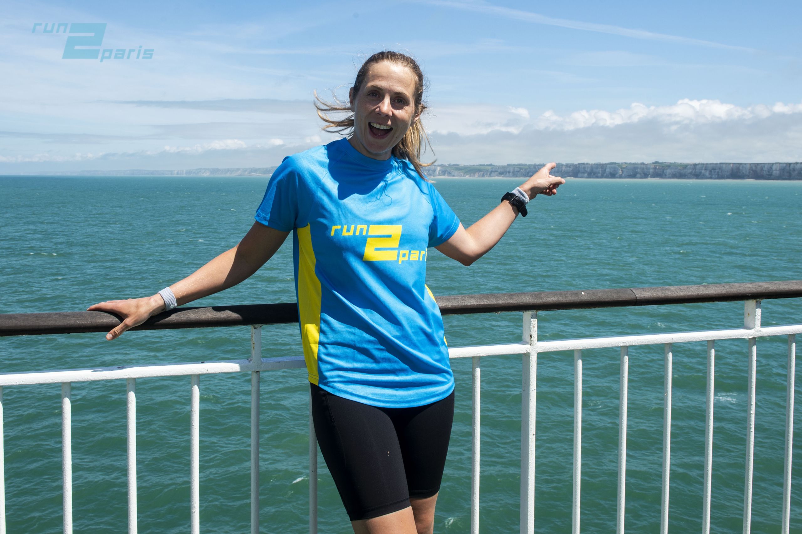 Bright woman in running gear smiling on ferry with sea and cliffs behind.