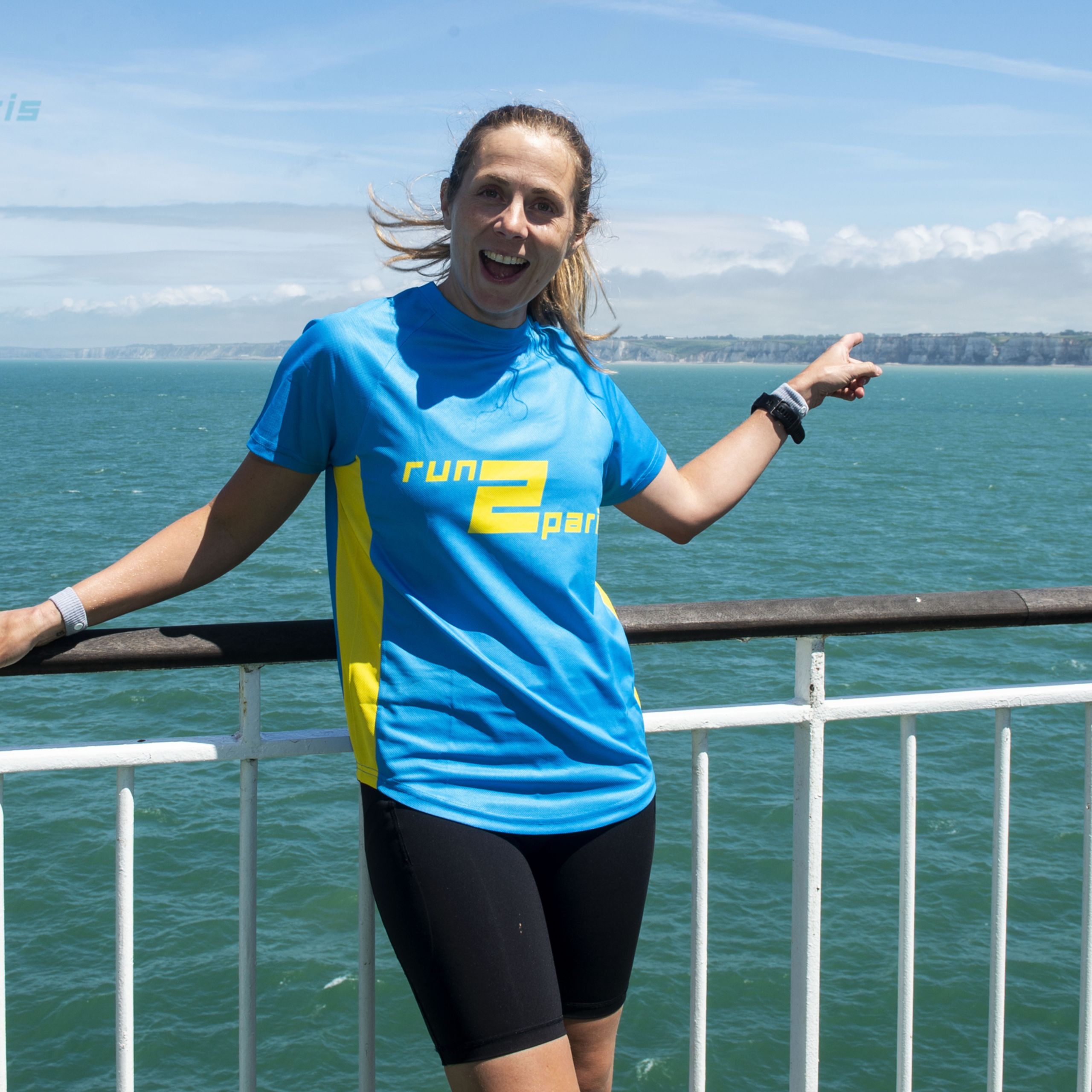 Bright woman in running gear smiling on ferry with sea and cliffs behind.