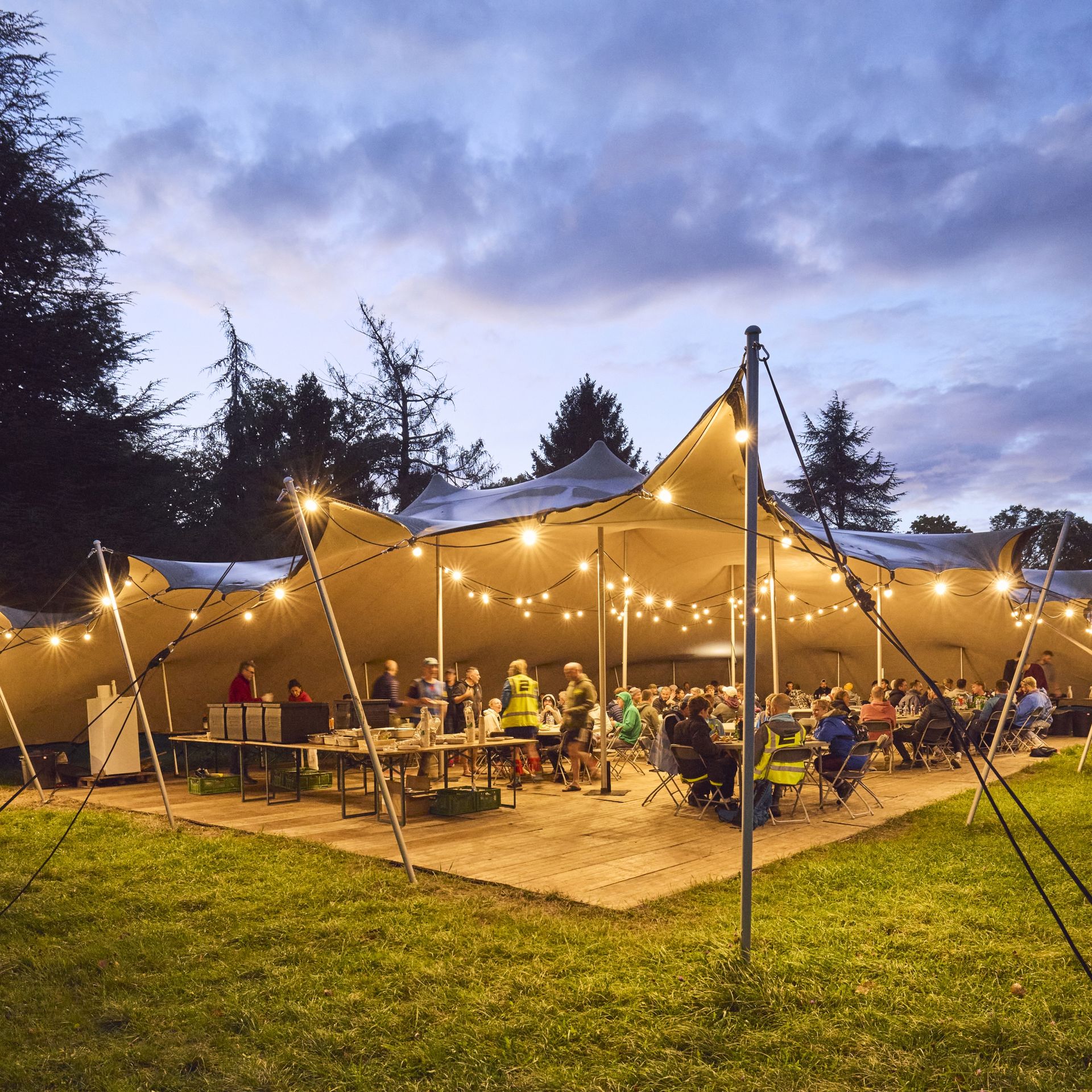 Vibrant evening at a relay race event with people under a large illuminated tent in a scenic outdoor setting.