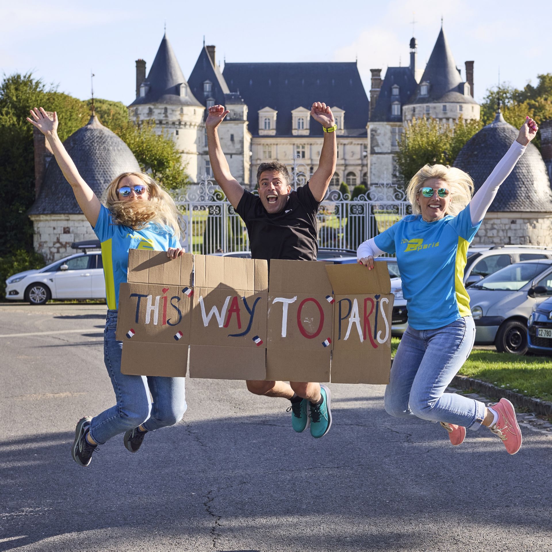 Energetic runners celebrating with a "This Way to Paris" sign in front of a castle during the relay run.