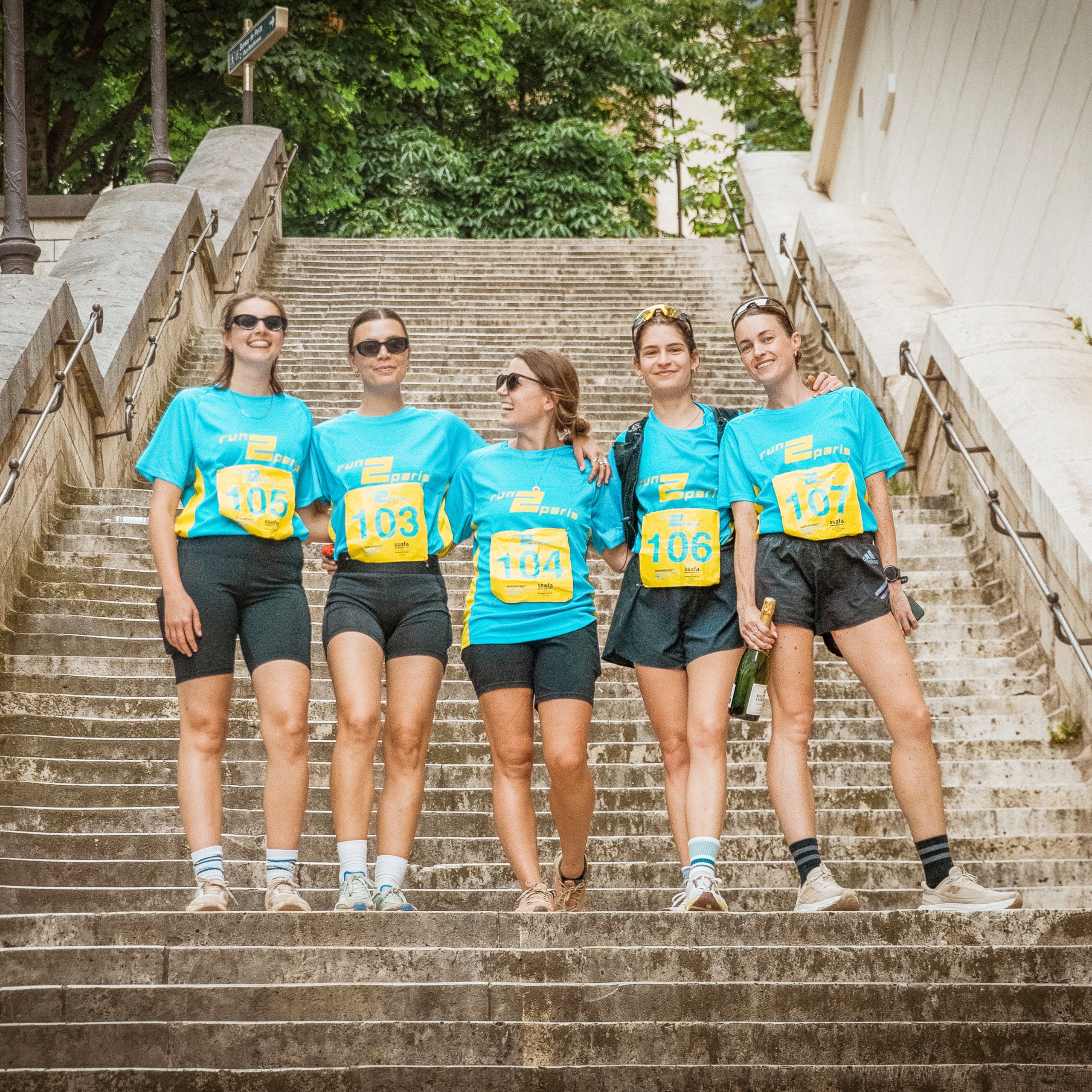 Bright female runners in blue event shirts standing on a staircase in Paris, celebrating during the Run2Paris relay race.
